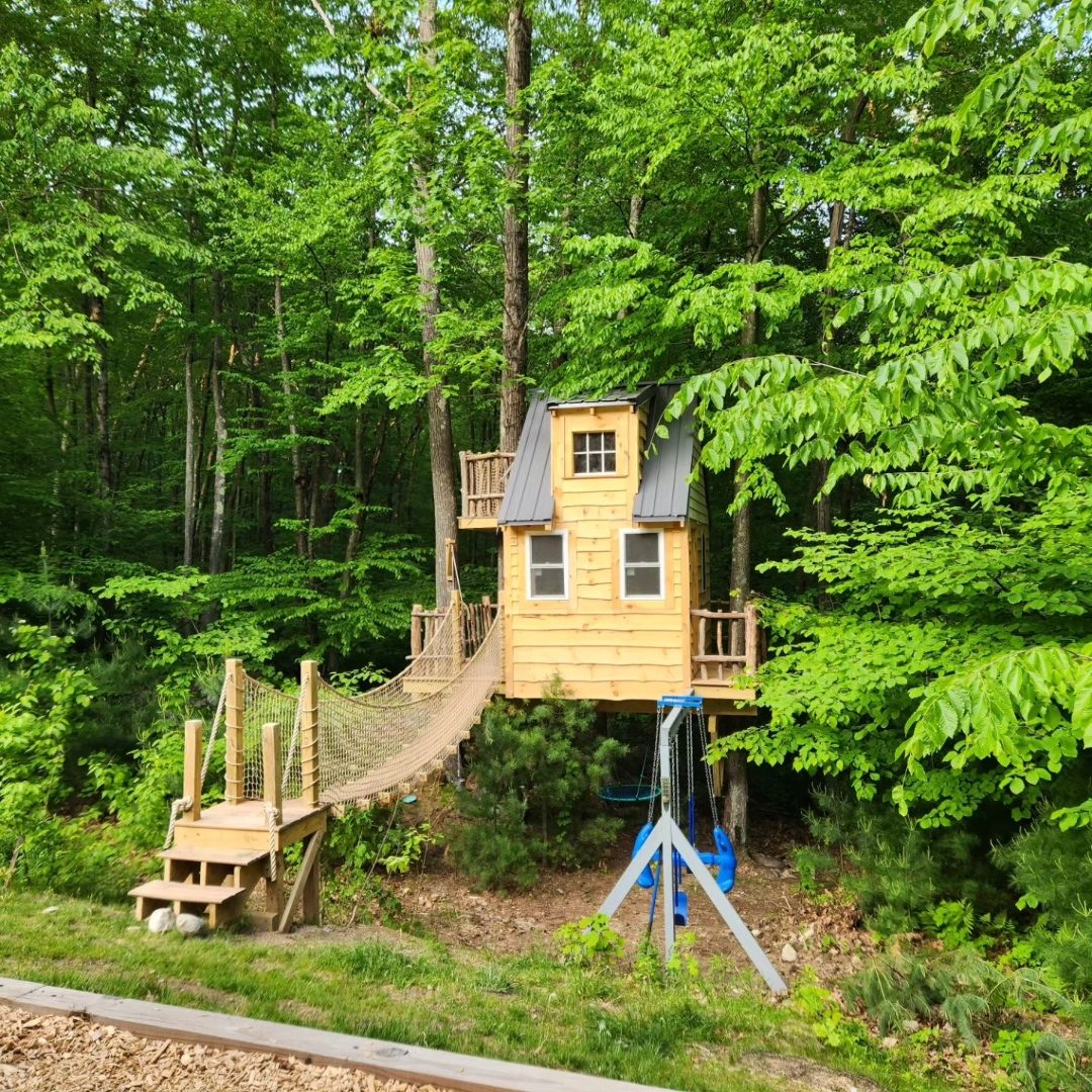 Custom treehouse with dormer, stairs, and wooded rope bridge setting