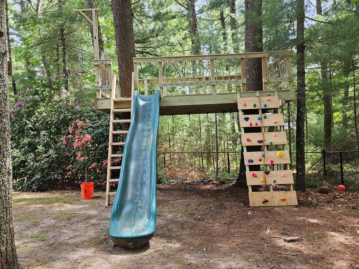 Backyard treehouse play area with blue slide, rock wall, and rope railing